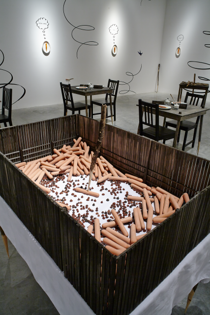 View of inside the wooden cage. There are approximately 30 hot dogs huddled together forming a circle around a single hotdog pierced by a wooden stake. There are small brown droppings under the hot dogs.     