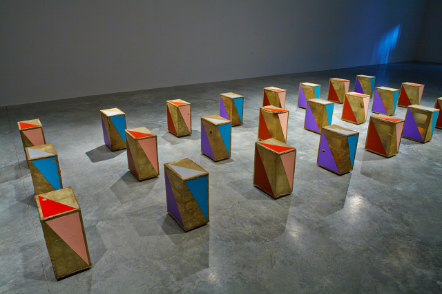 Wide angle of the installation. Three rows of eight box like objects stand vertically. The objects have brown base with different colours painted geometrically on t the object. 