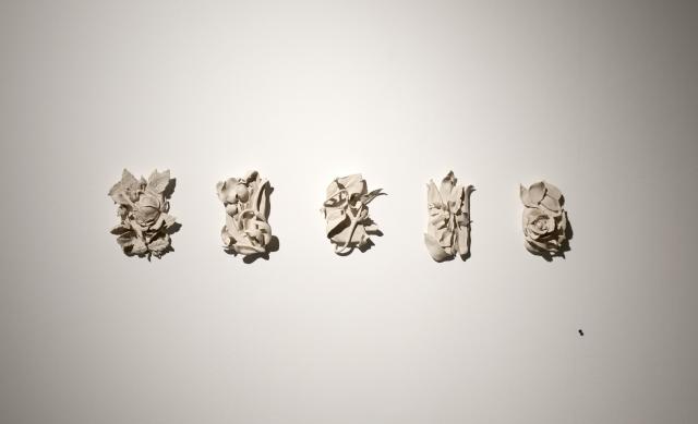 Detail of Hanging Garden 1-5 (2011-12). There are five unglazed beige ceramic objects. They each take the form of a different flower. Each form appears to be squished into a rectangle shape. 