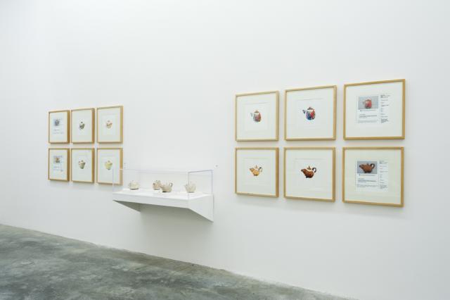 Twelve plywood framed prints of colourful teapots sit on the wall. In the center of the prints is a display case of said teapots. 