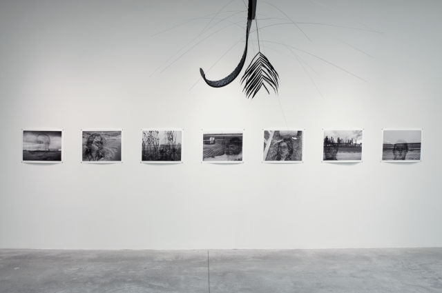 Image of Seeking the Human in an Inhumane Environment (2013). There are seven black and white photographs lined in a row. The sculpture Slavery? (2013) peaks in from out of frame. 