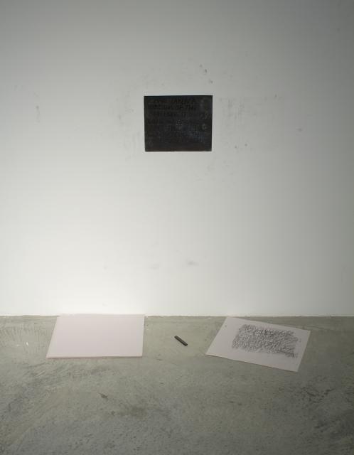 Metal block with text is hung on the wall. Two sheets of paper sit bellow it with a piece of charcoal. on closer inspection they are rubbings of the above metal casting. 