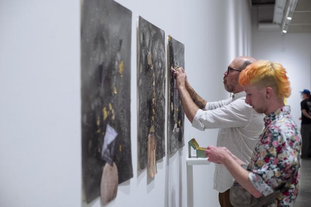 Detail of gallery attendees offering gold leaf to a water colour triptych . Heather Hart. New Numinous Negro: Mildred Lewis I,II,III. 2022  Ink, collage, mineral, gold leaf, participation on paper. Photo Credit to Elyse Bouvier.  