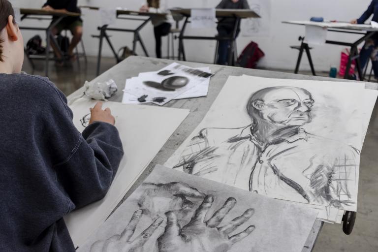 Teen faces + figures student working at their desk with images of their projects including a drawing of a persons torso and a hand.