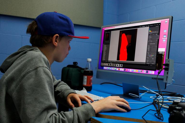 Teen photo student works at a computer in a tech lab with blue walls editing a photo in photoshop