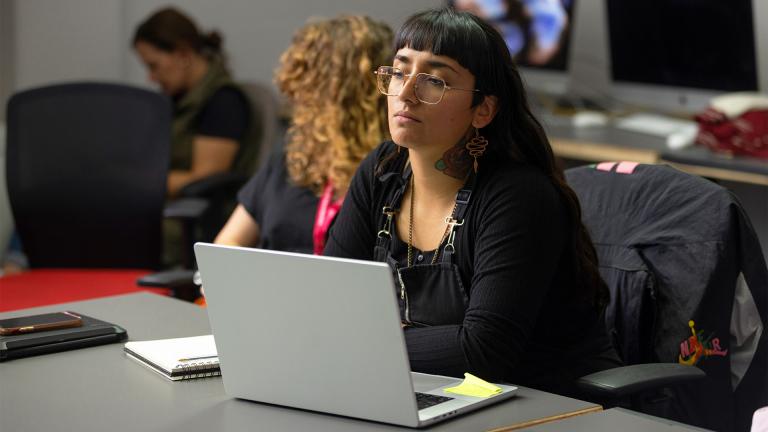 A focused student in a design classroom, working on a laptop with a notebook and sticky notes nearby. The modern learning environment features computers and engaged peers in the background, highlighting a collaborative and creative atmosphere.