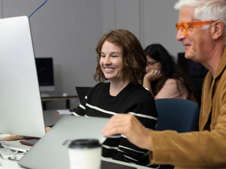 Design students collaborating in a digital lab, engaging with creative projects on desktop computers. The atmosphere is dynamic and interactive, with an instructor providing guidance and students exchanging ideas in a modern learning environment.