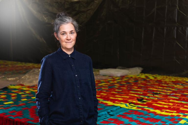 A person with hair tied up in a bun stands in front of a large-scale textile installation with a woven pattern in red, blue, and yellow. She wears a dark blue button-up shirt and has a calm, confident expression. The background features a dark, draped material adding depth to the scene.