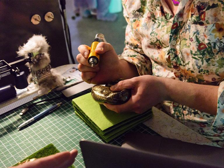 Close-up of an artist engaged in a hands-on creative project, using a precision tool to work on a small sculptural piece. The workspace is filled with materials, tools, and textures, highlighting an intricate and experimental artistic process.