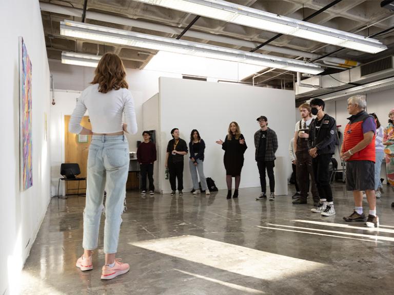 Art critique session in a bright, open gallery space, where a student presents their artwork to a group of peers and faculty. The group listens attentively, fostering a collaborative and supportive environment for artistic growth and discussion.