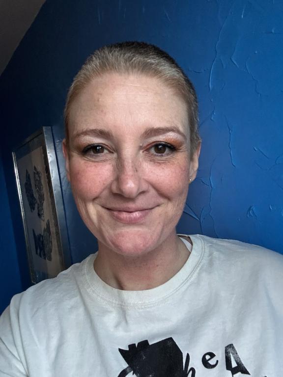 A smiling person with closely cropped hair wearing a white graphic T-shirt, standing in front of a textured blue wall, with a warm and friendly expression.