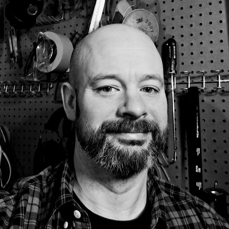 A black-and-white portrait of a bearded man with a shaved head, wearing a plaid shirt. He is smiling slightly and looking at the camera. The background features a pegboard wall with various tools, including tape rolls, screwdrivers, and measuring instruments.