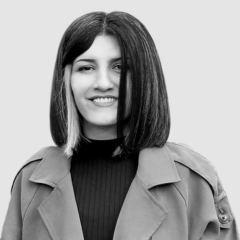 Black and white portrait of a woman with shoulder-length straight hair, featuring a distinctive blonde streak on one side. She is smiling subtly and wearing a dark turtleneck sweater under a structured coat. The background is a neutral gray, giving a clean and professional appearance.