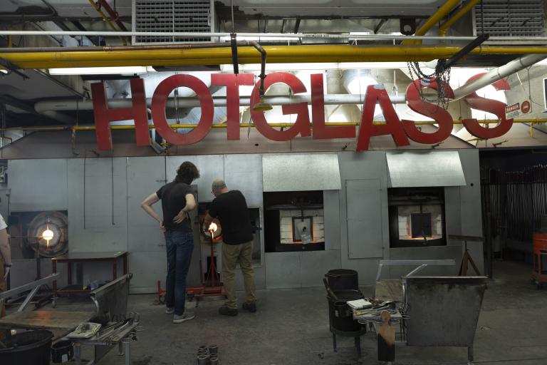 Glass Studio Two people working with furnaces in the AUArts glass studio under a large red “HOT GLASS” sign.