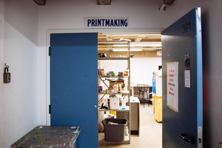 A blue door labeled “Printmaking” stands open, revealing shelves with inks, tools, and supplies inside a studio. A sign on the door reads, “Last one out shuts the door.