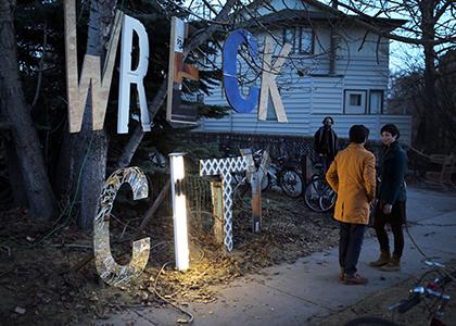 Large Wreck City Sign made of reclaimed wood and scrap, sitting near a house beside a sidewalk with event attendees chatting beside it