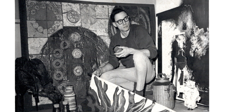 A black and white photo of a kneeling Dennis Evans showing student work at Gunther’s Mobilia Furniture Showroom, 1968