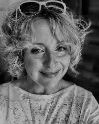 black and white photo of Jenifer Darbellay, seen from the shoulders up. She has short wavy blond hair curling around her face and she is wearing sunglasses on her head. She is smiling. The photo is a casual, relaxed shot, probably a selfie photo