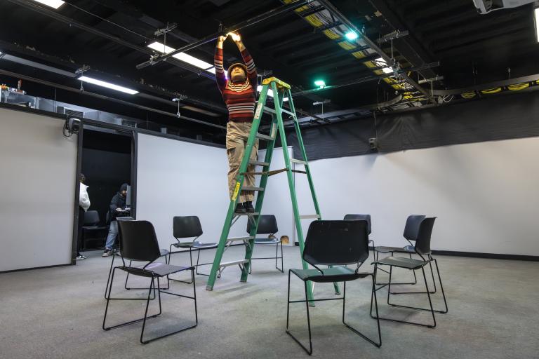 Person standing on a ladder adjusting overhead equipment in a studio space, with chairs arranged in a circle below.