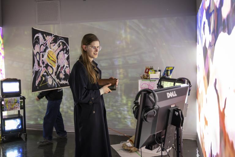 Contemporary media art installation featuring a person interacting with projected visuals and monitors in a gallery space, surrounded by abstract artwork and video screens.